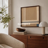 A Collected Morning cream, toasted sand, and indigo yarn wall art displayed above a wood cabinet beside a curved cream chair in a reading corner.