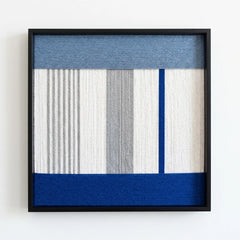 Professional studio shot of the Absolute Zero hand-woven wool wall art featuring blue and grey geometric patterns in a black frame.