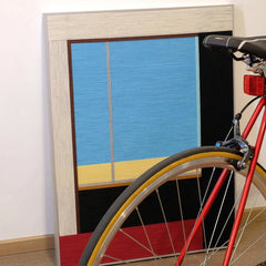 After the Beach Walk yarn wall art displayed leaning against a bicycle wheel, emphasizing its vibrant blue, gold, and red stripes