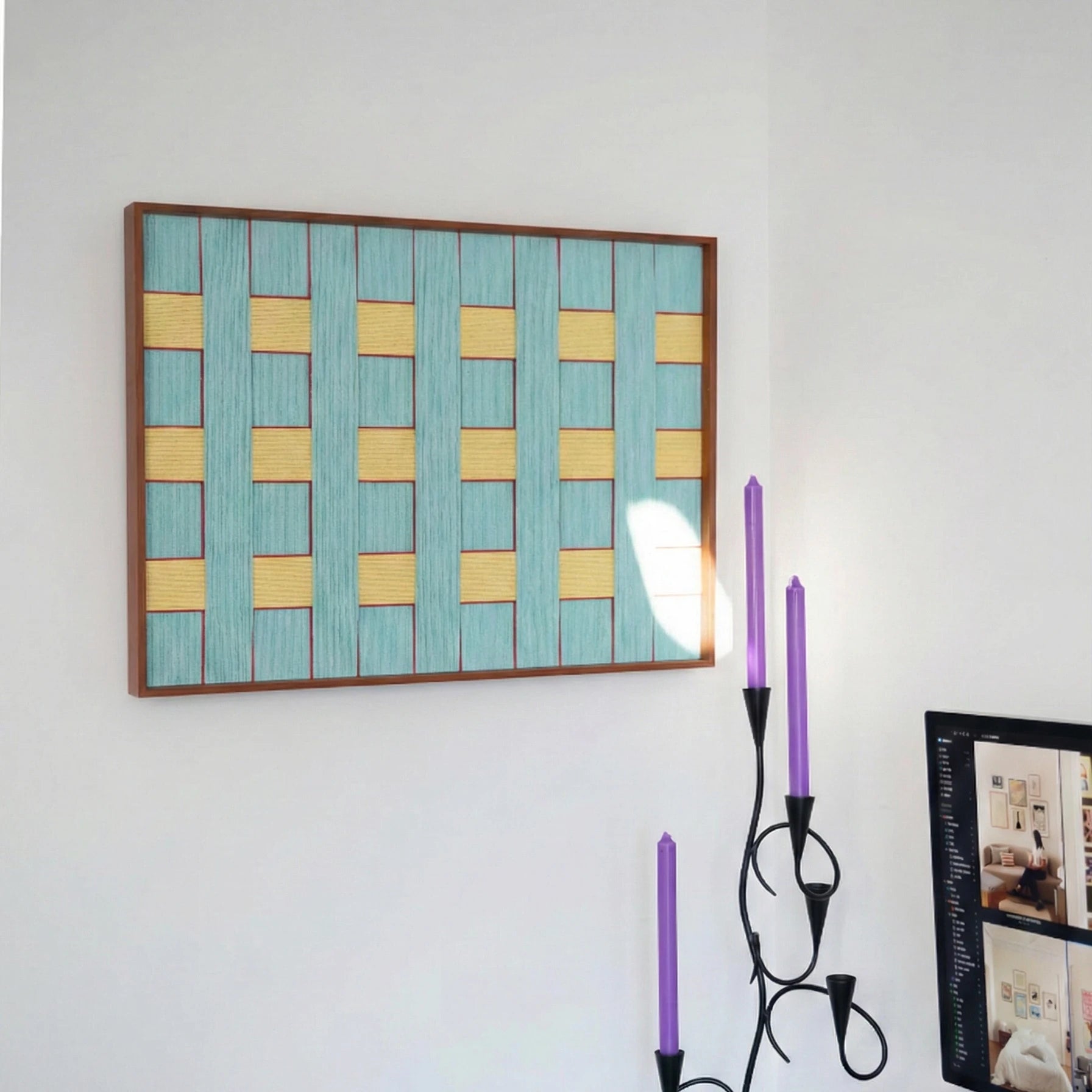 Horizontal textured wool wall art in a wooden frame placed on a white wall above a decorative candelabra with purple candles.