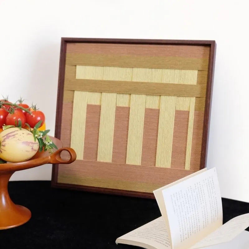 Autumn Mountain hand-woven wool yarn wall art featuring alternating pale gold and taupe vertical stripe panels in a natural wood frame, displayed on a black tabletop.