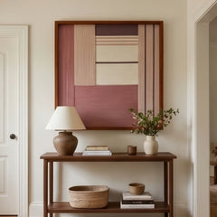 Striking square hand-woven wool tapestry positioned perfectly above a dark wooden console table adorned with a small lamp and ceramics in a bright hallway.