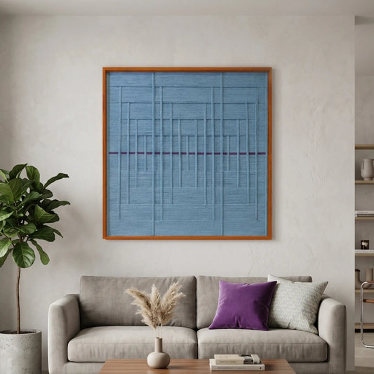 Square hand-woven wool wall art in a cherry wood frame featuring a monochromatic slate blue textured grid pattern with a single horizontal purple accent line, styled above a grey sofa.