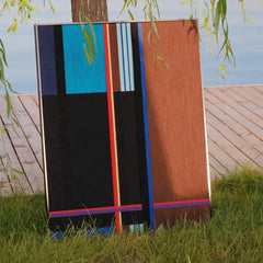 Blue Tide Night Run yarn wall art placed on a grassy riverbank, emphasizing its outdoor-friendly wool texture and vibrant stripe contrast.