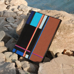 Blue Tide Night Run yarn wall art resting on a rocky shoreline, highlighting the sturdy frame and bold stripe composition against natural stone.