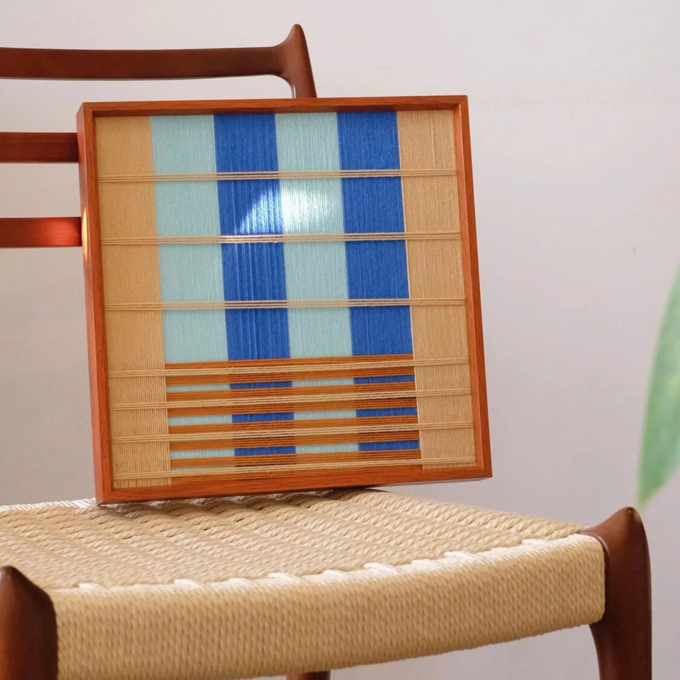 Chuan yarn wall art leaning on a chair, emphasizing compact square proportions and bold blue-stripe composition