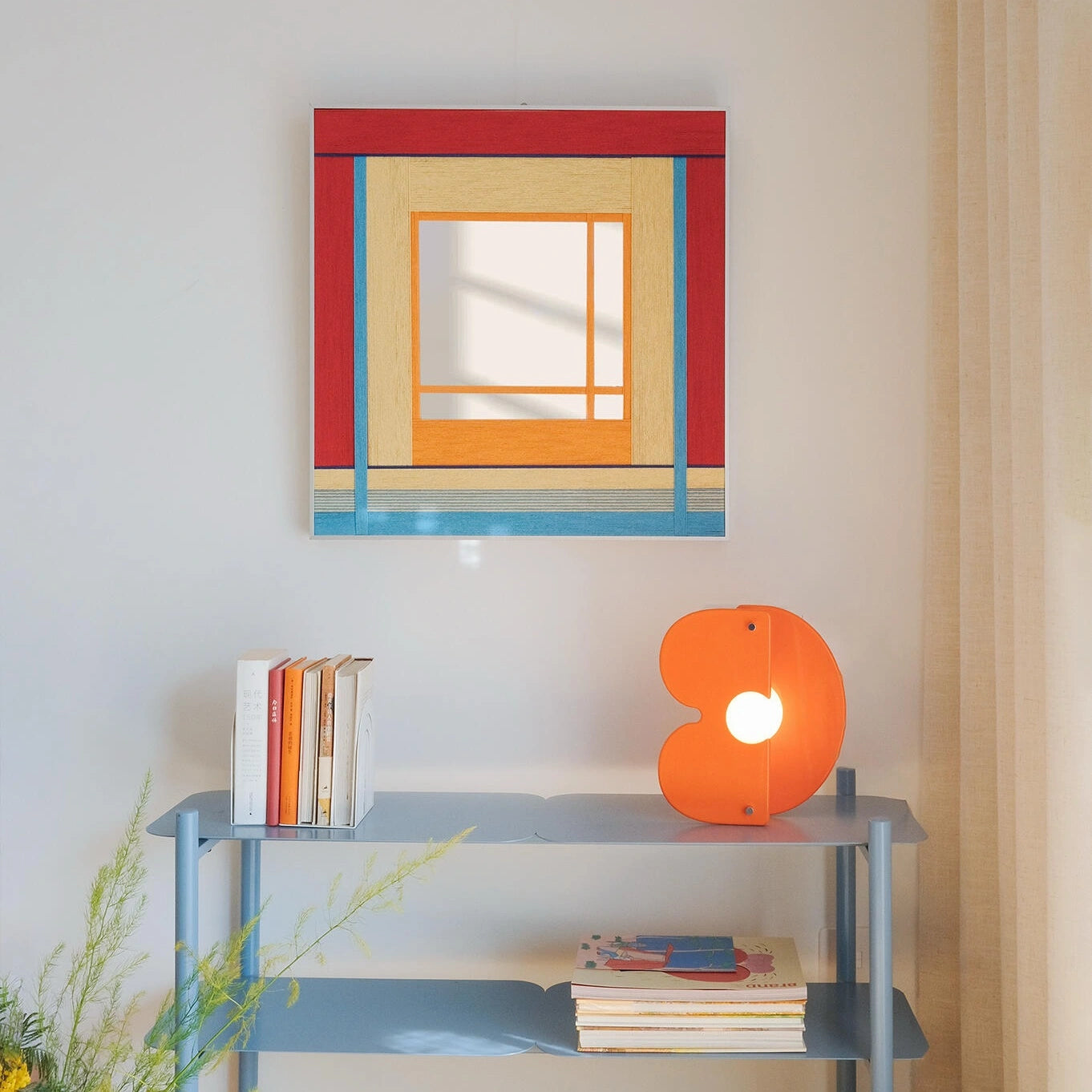 Framed fiber art mirror titled Fields of Blossoms featuring a geometric red, yellow, and blue design, styled above a blue shelving unit.