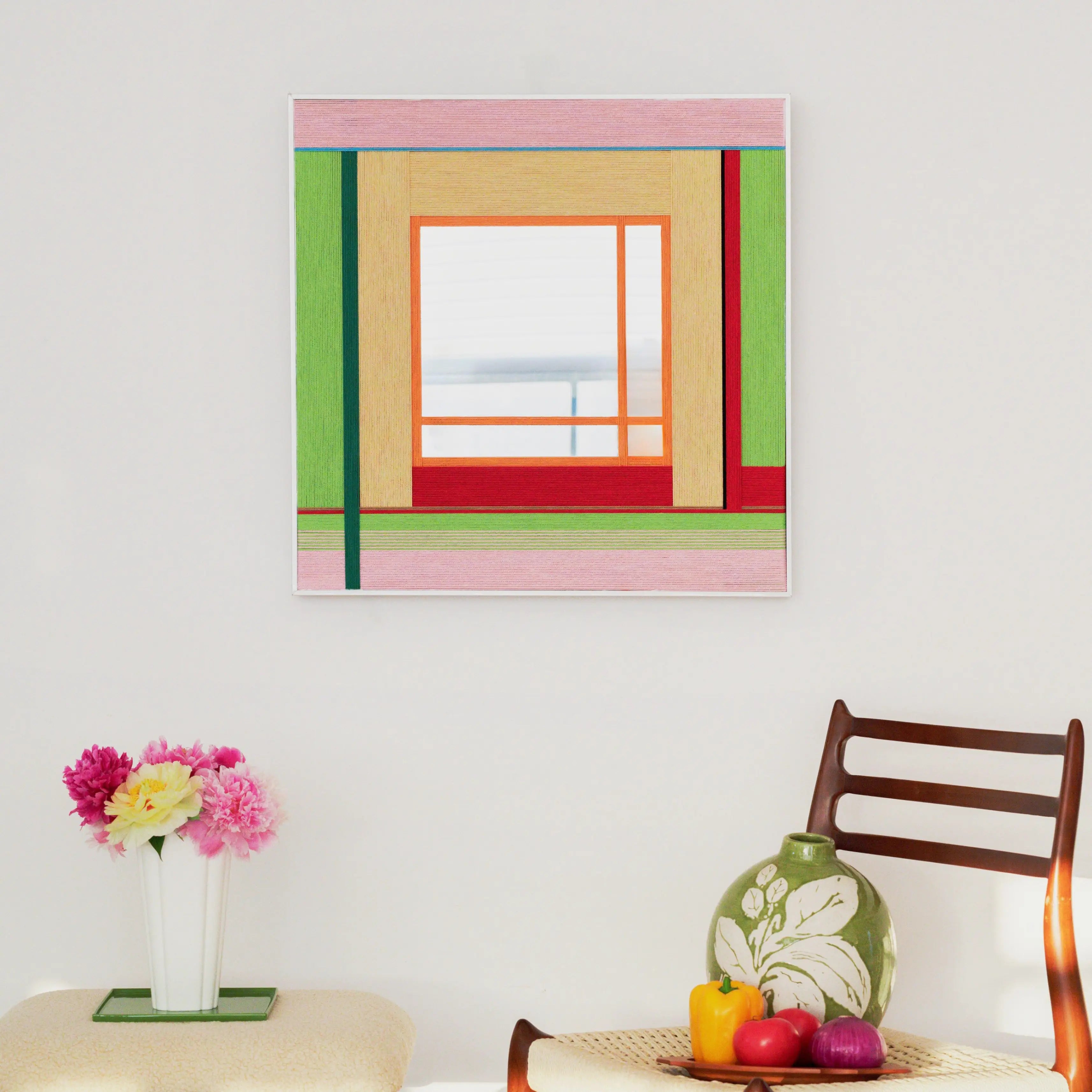 The Olive Tree Romance woven mirror hanging on a white wall above a chair, adding a soft pop of color to a minimalist living space.