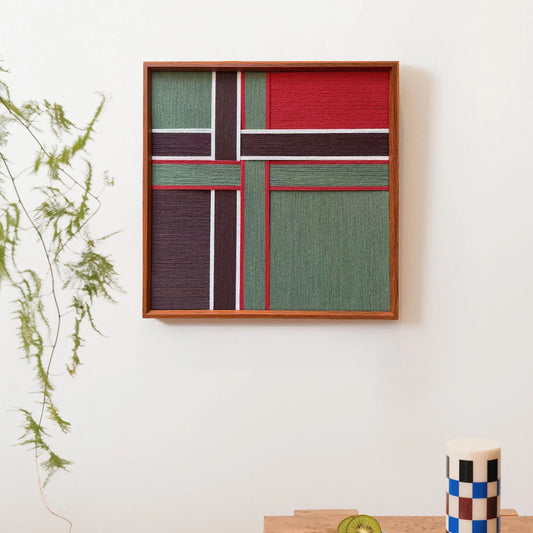 Framed hand-woven wool art featuring geometric red and green blocks with white lines resembling a gift ribbon, displayed on a white wall.