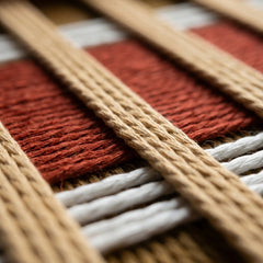Extreme low-angle macro shot of the Gilded Coronation hand-woven textile art lying flat, highlighting the textured rust-red and mustard-gold vertical yarn stripes.