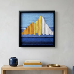 Square hand-woven wool wall art in a black frame depicting an abstract mountain landscape with golden and white peaks against a blue sky, hanging above a light wood console table.