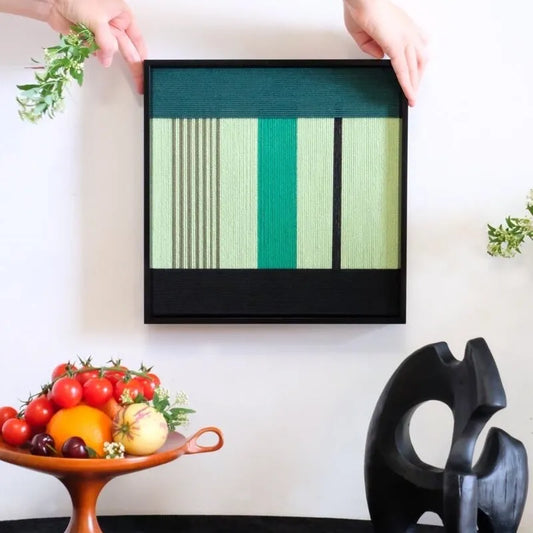 Green Branches hand-woven yarn wall art in alternating emerald and mint green vertical stripes, framed in sleek black wood and displayed above a console table.