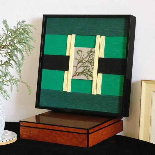 Green Glow hand-woven wool yarn wall art featuring alternating emerald and forest green vertical stripes framing a central cream botanical panel, displayed on a console table with decorative plant