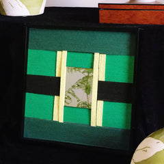 Hand-held view of Green Glow yarn wall art showing its compact square form, black wood frame, and precise stripe pattern of emerald and forest green
