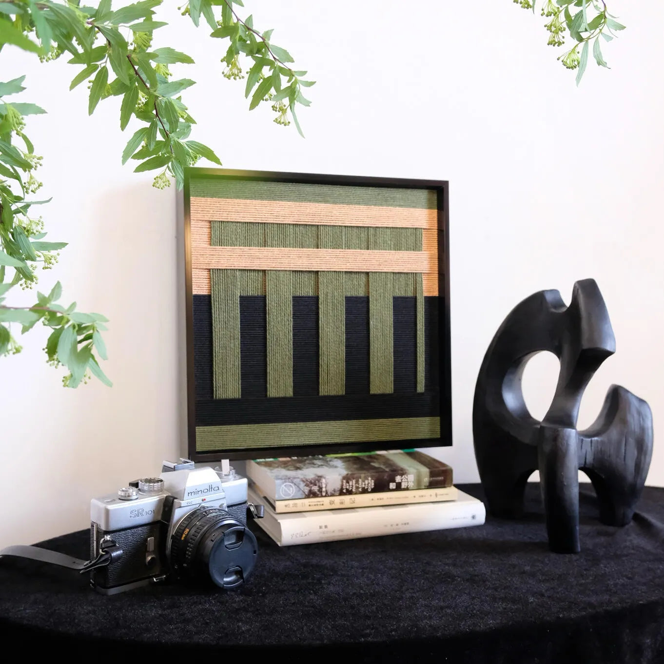 The Serene Wilderness fiber art piece displayed on a black table with vintage cameras and books, showcasing moody interior styling.