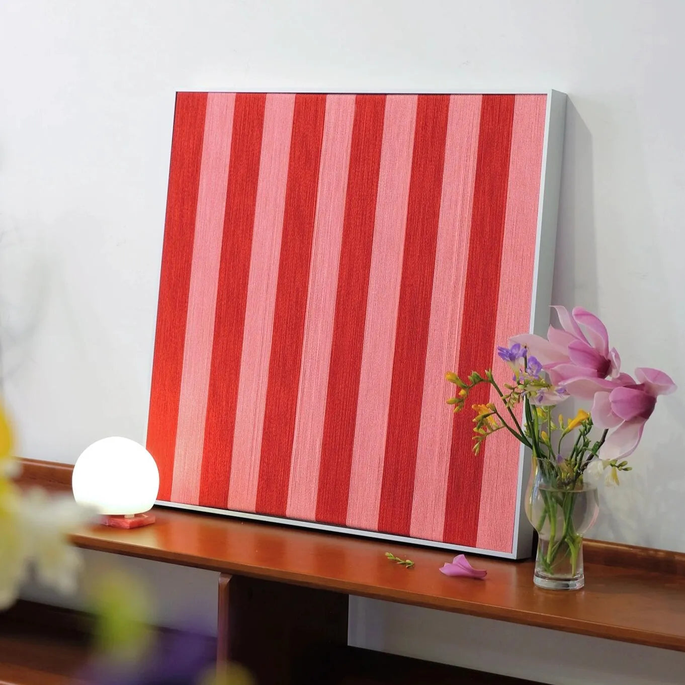 Angled view of Hana & Alice yarn woven art leaning on a tabletop beside a lamp and vase, showcasing the handcrafted texture and depth of the red stripe pattern