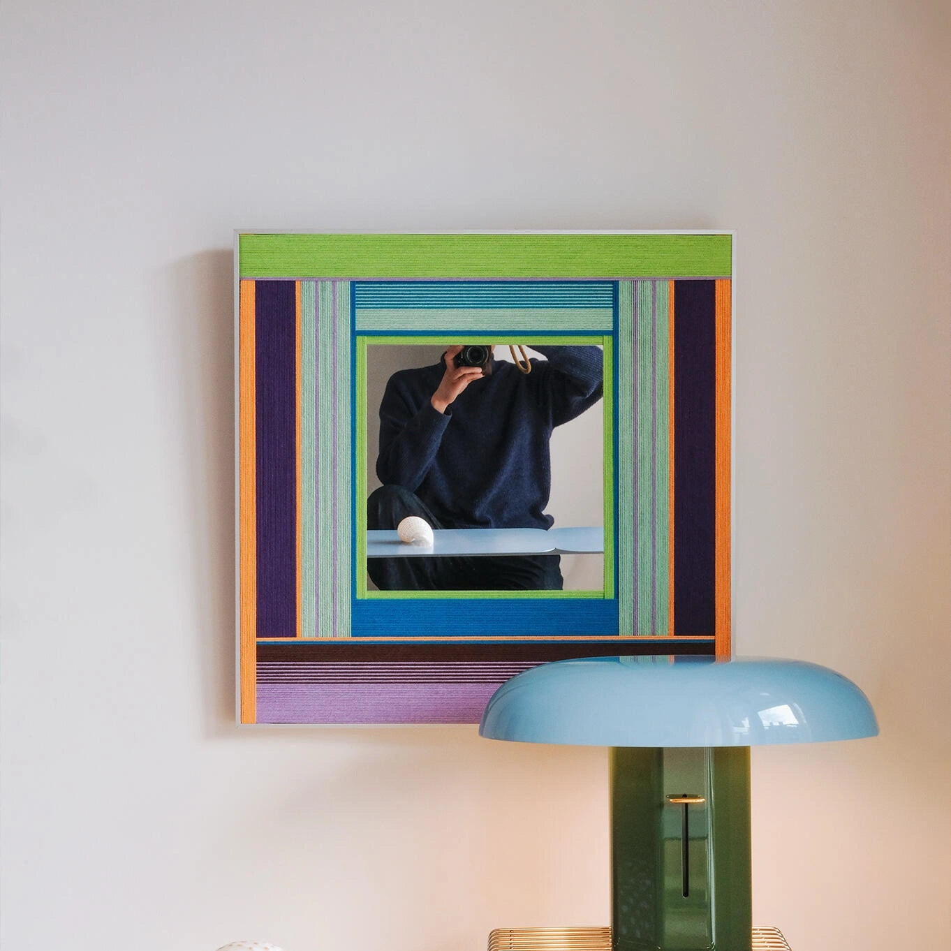 Hidden Forest hand‑woven yarn mirror wall art with alternating moss green, lavender, and natural wood stripe borders framing a central mirror, displayed above a desk with a teal lamp.