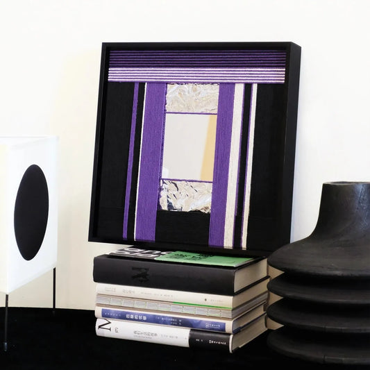 HOUTIAN hand-woven wool yarn mirror wall art with alternating purple and black vertical stripes framing a central mirror panel, displayed on a console table between speaker and books.