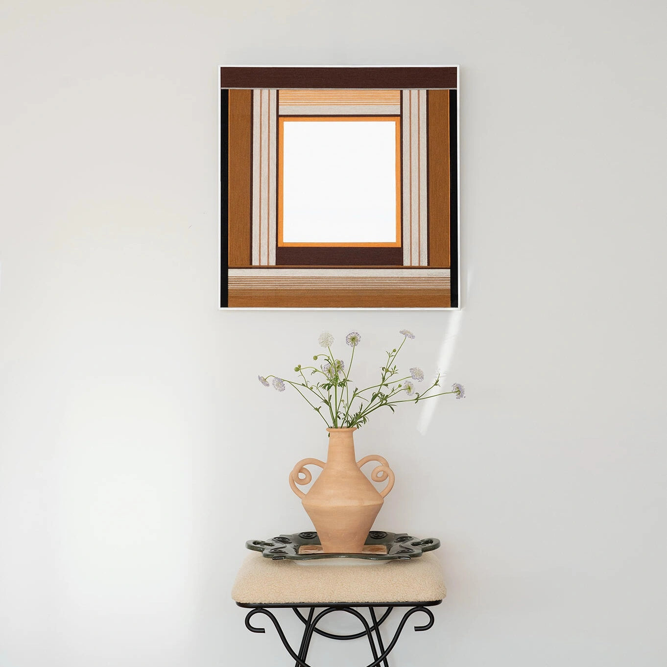 The hand-woven fiber mirror hanging above a small console table with a vase, adding a touch of handcrafted texture to a minimalist white wall.
