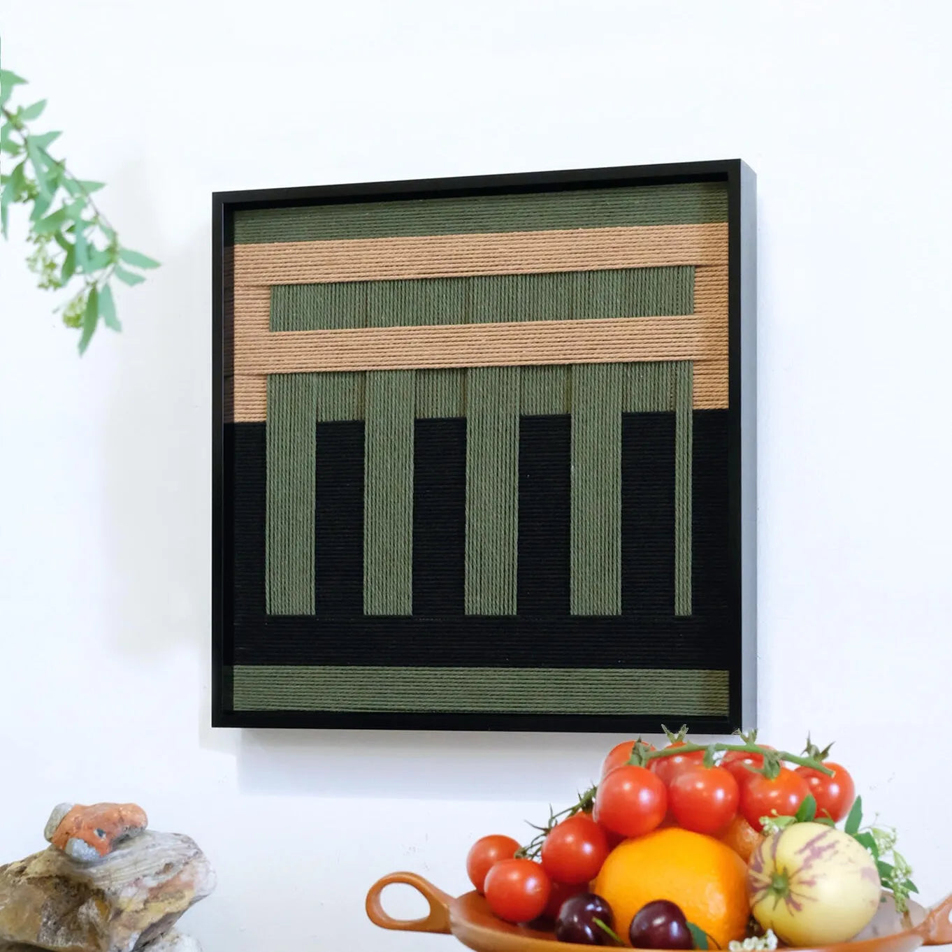The geometric fiber art hanging on a white wall above a fruit bowl, adding a natural and serene touch to a modern dining space.