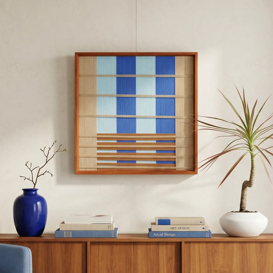 Square hand-woven wool wall art in a cherry wood frame featuring vertical stripes of cobalt and sky blue on a tan background with horizontal wood slat accents, styled above a wooden credenza.