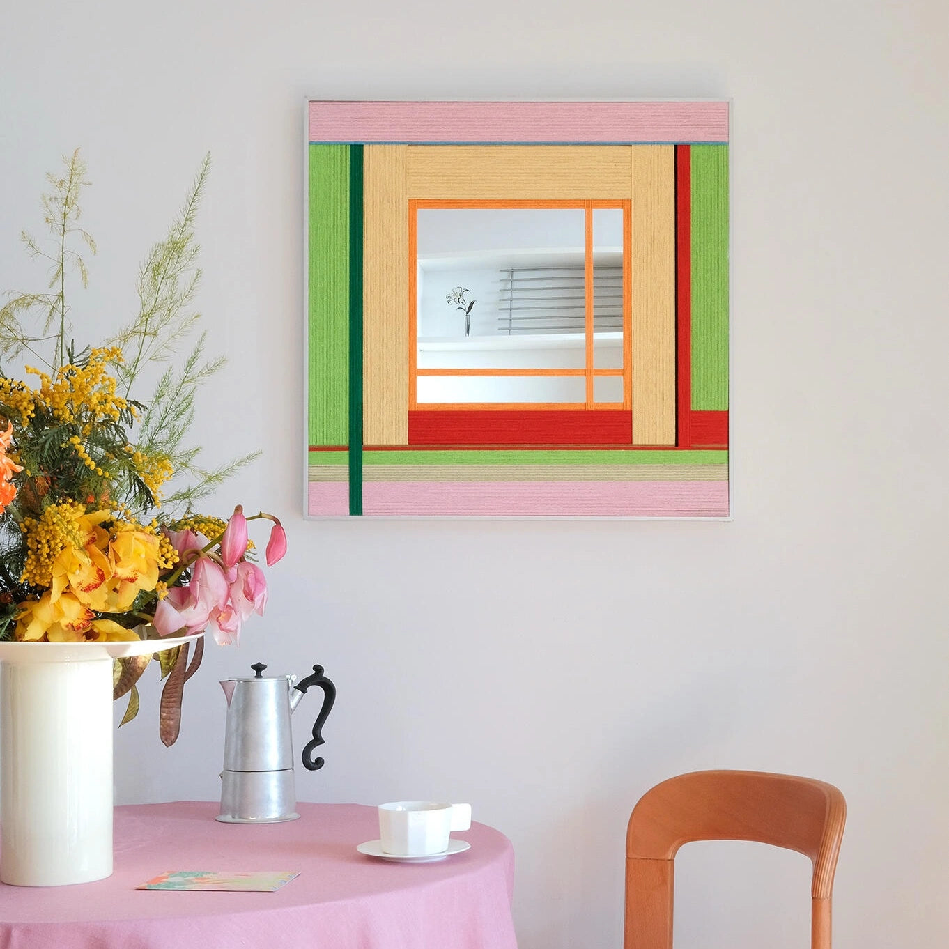 Framed fiber art mirror titled Olive Tree Romance featuring pink and green woven wool, displayed above a pink dining table with flowers.