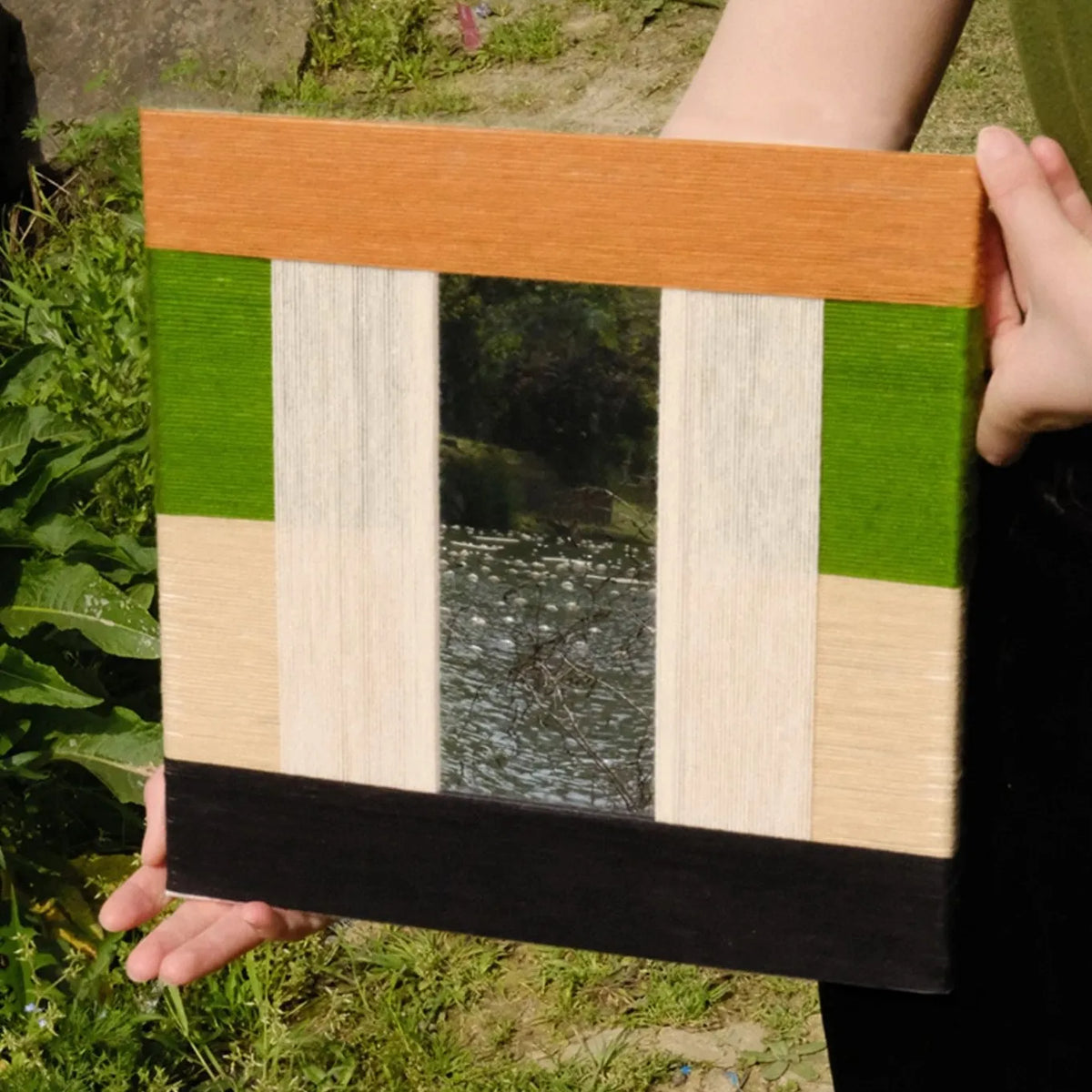 Hand-held view of Pastoral Elegance yarn wall art showing its exact square proportions and tight weave of green and beige panels