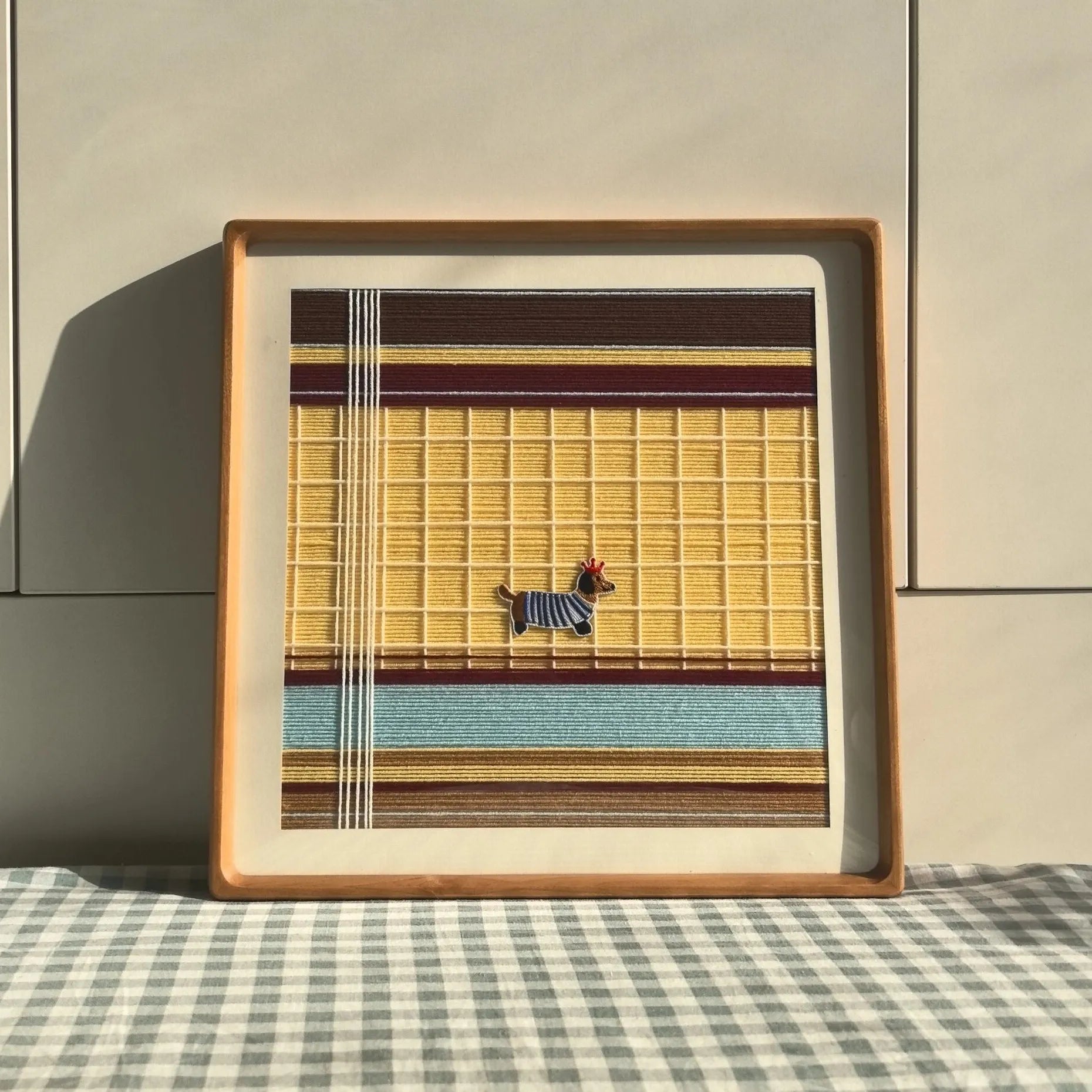 Pawsy Puppy hand-woven yarn wall art featuring a small black-and-white puppy silhouette centered on mustard yellow grid stripes with aqua and white accents, framed in natural wood and displayed on a gingham tabletop.