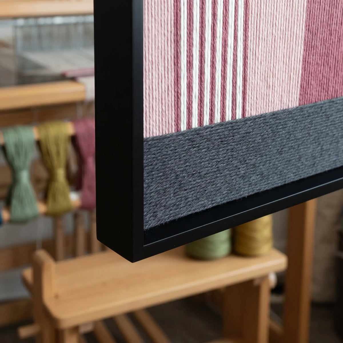 Side detail of the framed fiber art displayed in a workshop setting with a weaving loom and yarn in the background.