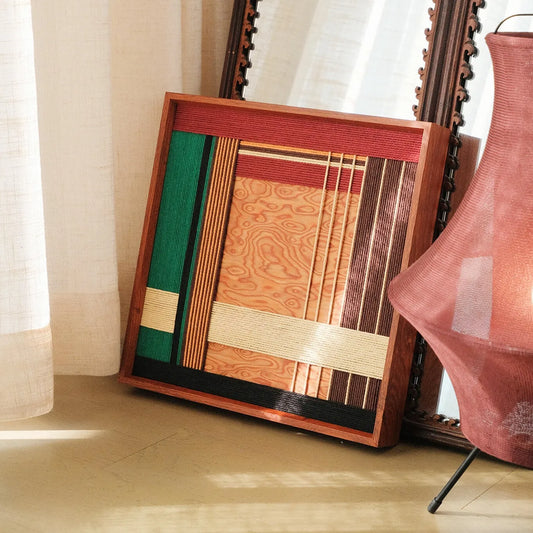 Saint Christmas hand-woven wool yarn wall art with alternating festive red, emerald green, and natural wood-tone stripes in a dark wood frame, displayed on a console table beside a violin and decorative plant