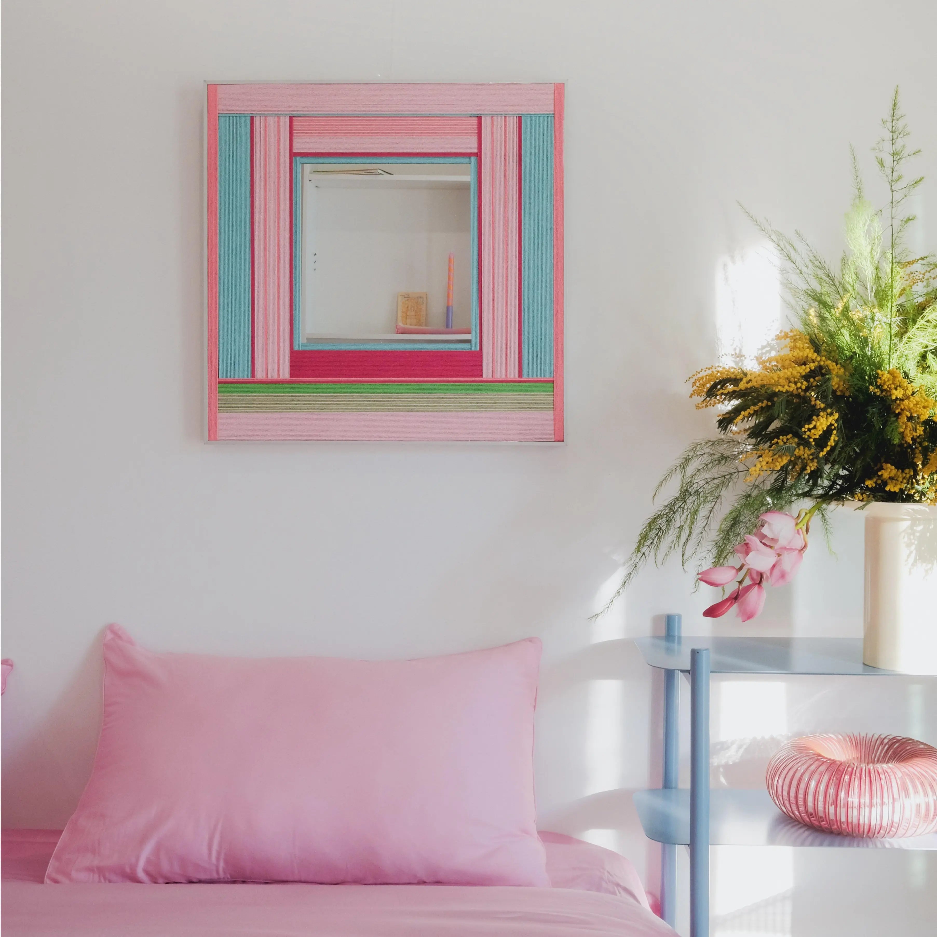 Secret Crush yarn mirror art styled on a shelf beside greenery and a pink cushion, showcasing its compact square form and fresh stripe palette.