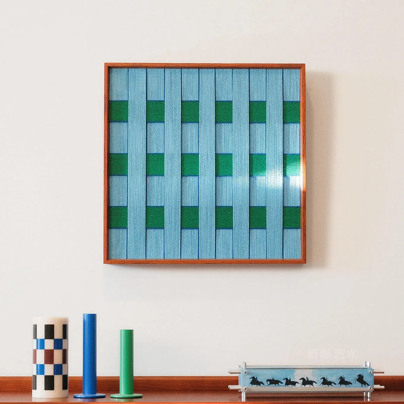 Seine Riverbank hand-woven wool yarn wall art with alternating green-blue vertical stripes in a natural wood frame, displayed on a console shelf with minimal décor