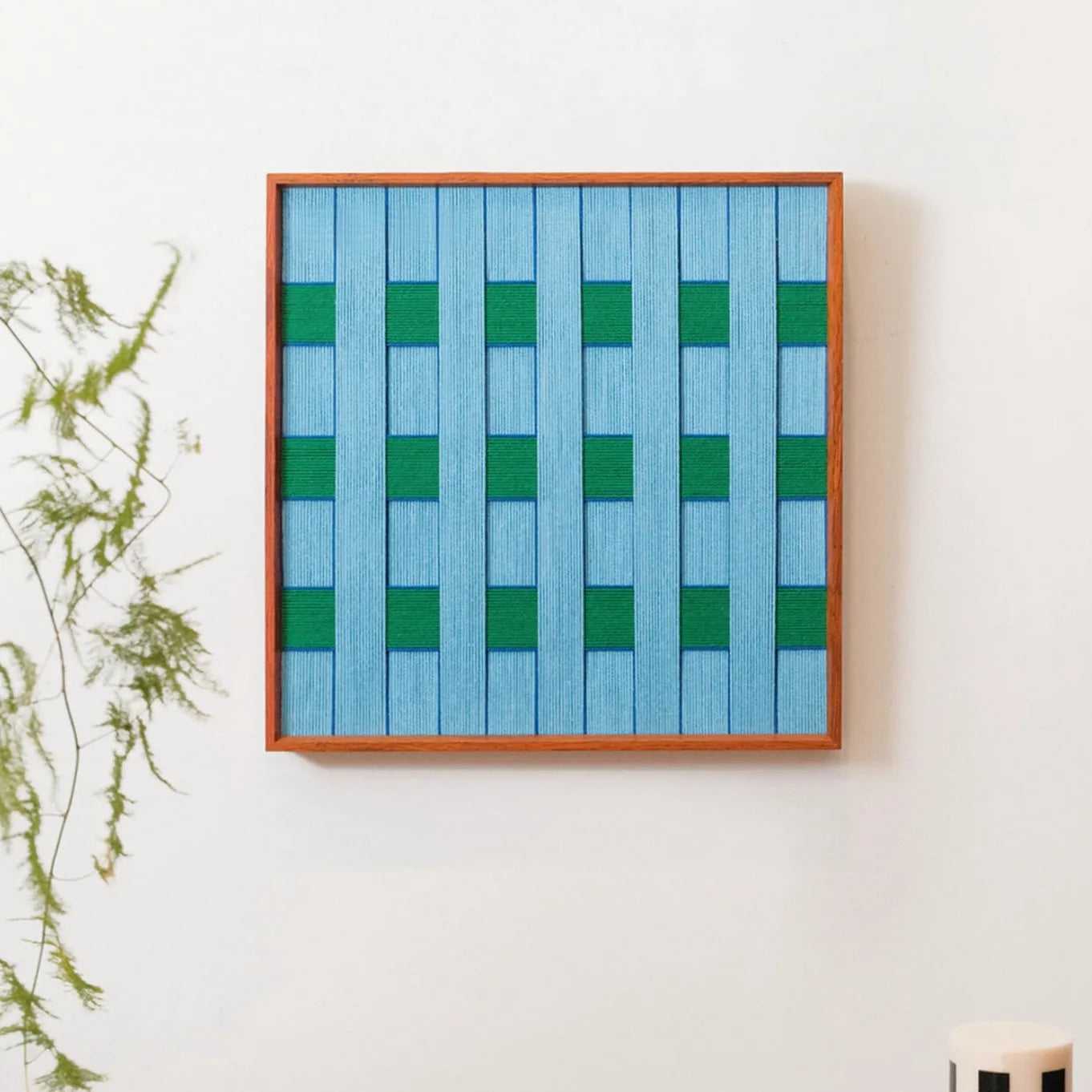 Seine Riverbank yarn wall art mounted on a white wall next to a potted plant, highlighting its vivid green-blue stripe pattern