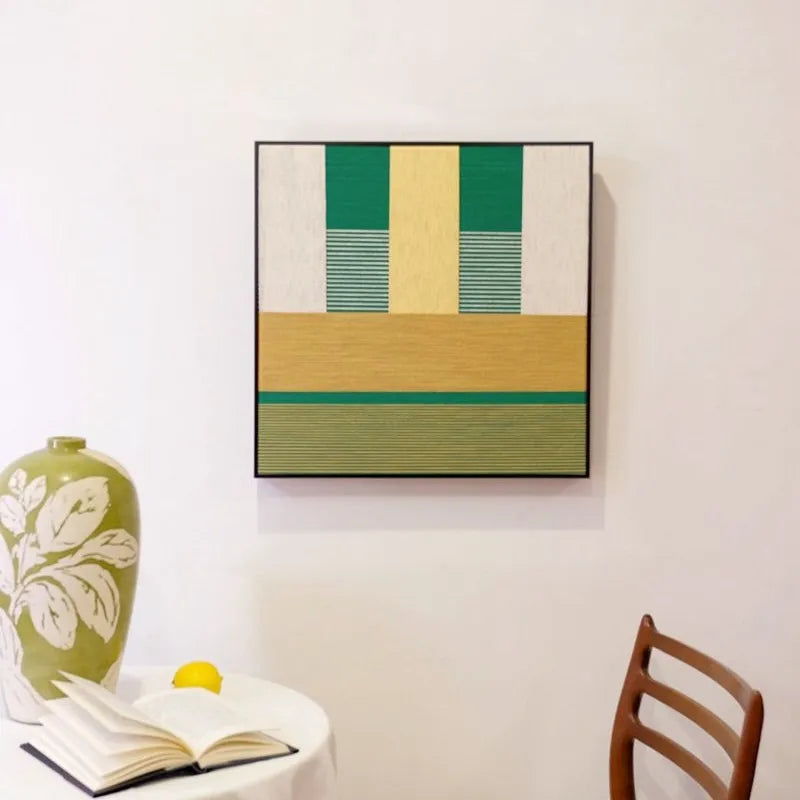 Spring Breeze Wheat Field yarn wall art hung on a wall above a chair, emphasizing its exact square proportions and clean geometric stripe design