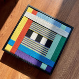 Sunlit angled flat lay of Childhood Old Toys SQ005 showing colorful yarn blocks and a slim black frame on a wood floor
