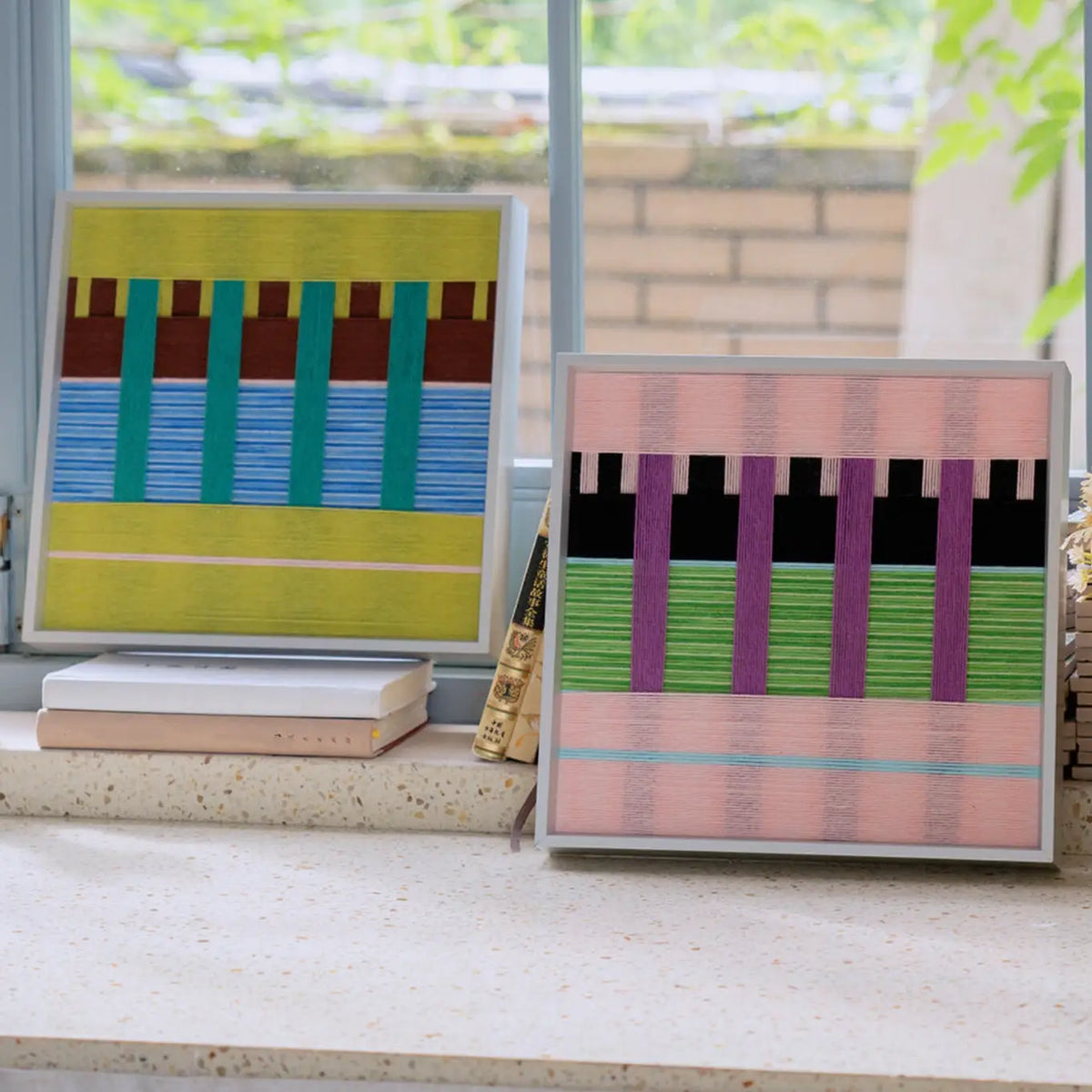 Woodland Mirror and Pastel Nocturne SQ116 textile artworks displayed together on stacked books near a bright window.