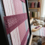 Angled close-up of Merlot Veil SQ133 showing the black frame and dimensional burgundy bands over pink and grey yarn.