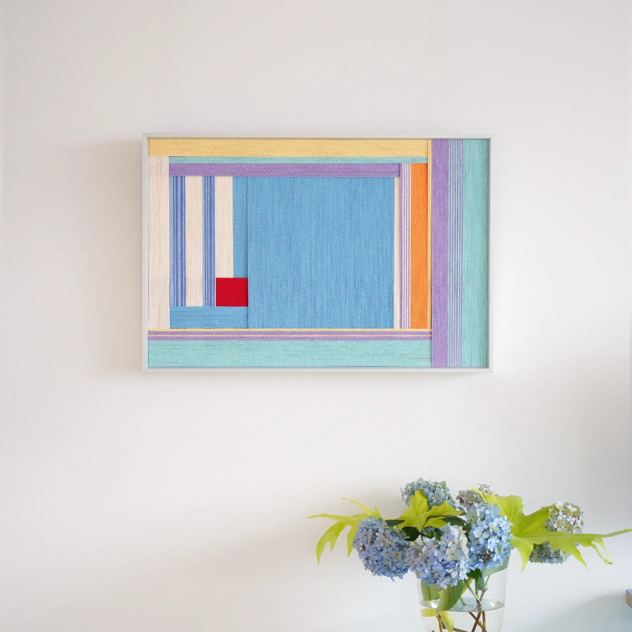 Hand-woven wool wall art with vibrant blue, orange, and purple geometric blocks hanging above a vase of blue hydrangeas.