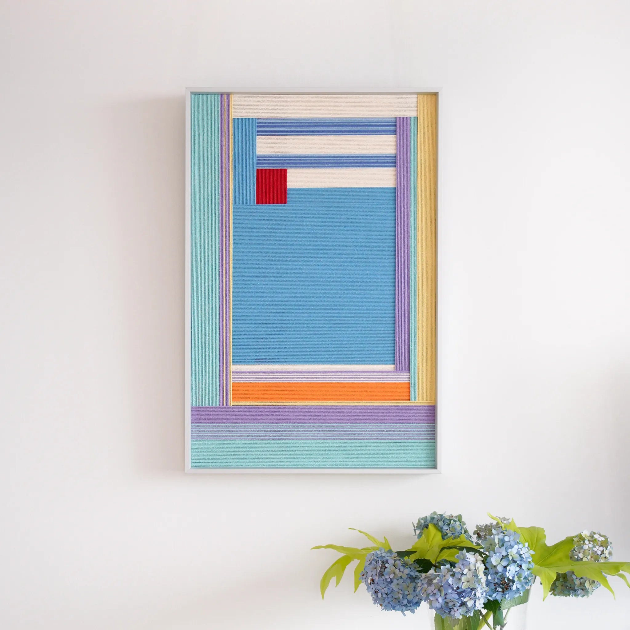 A portrait-oriented framed fiber art tapestry featuring colorful abstract wool weaving displayed on a clean white wall with flowers.