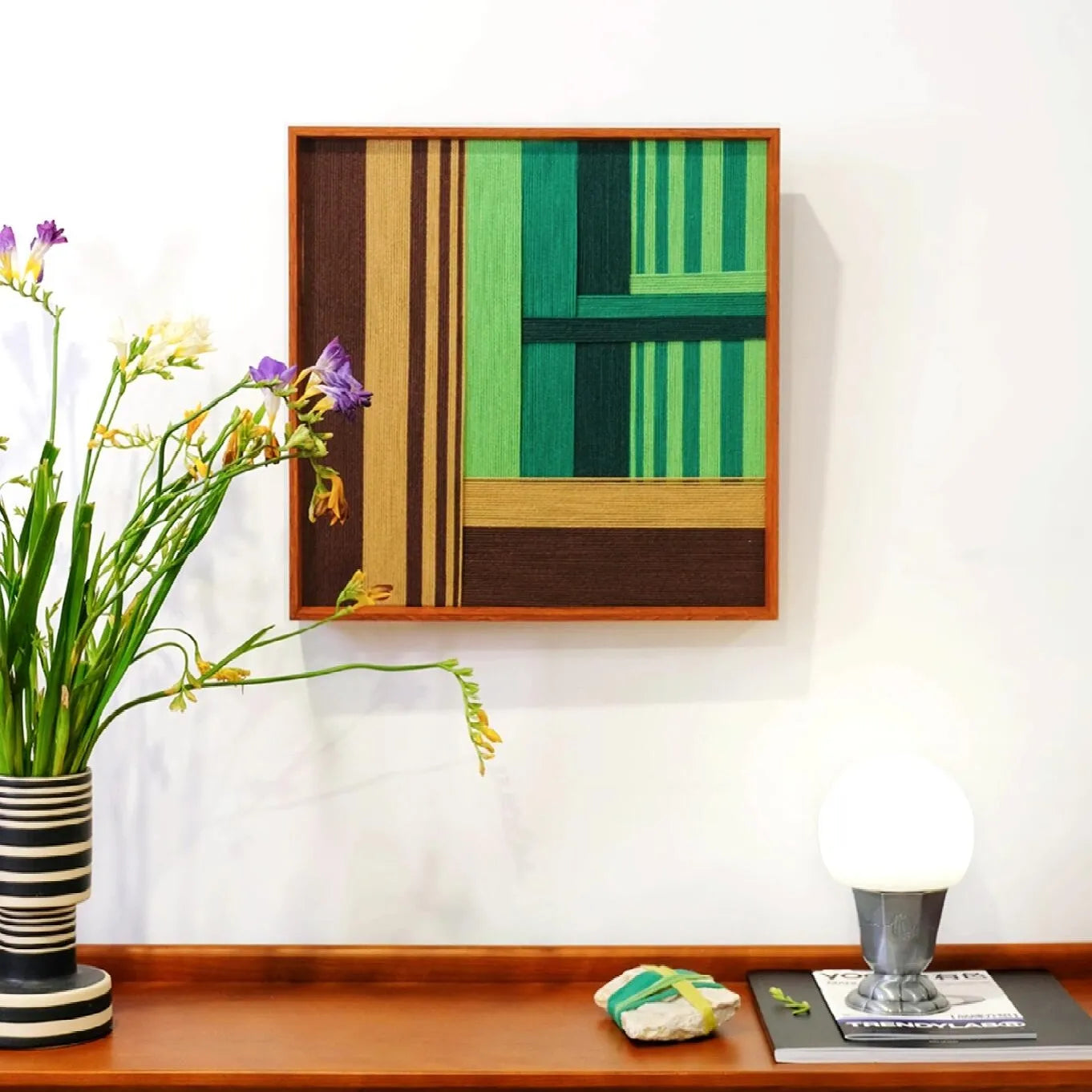 Framed "Summer Mountain Walk" hand-woven wall art featuring green and brown geometric blocks, styled on a wooden sideboard with fresh yellow flowers.