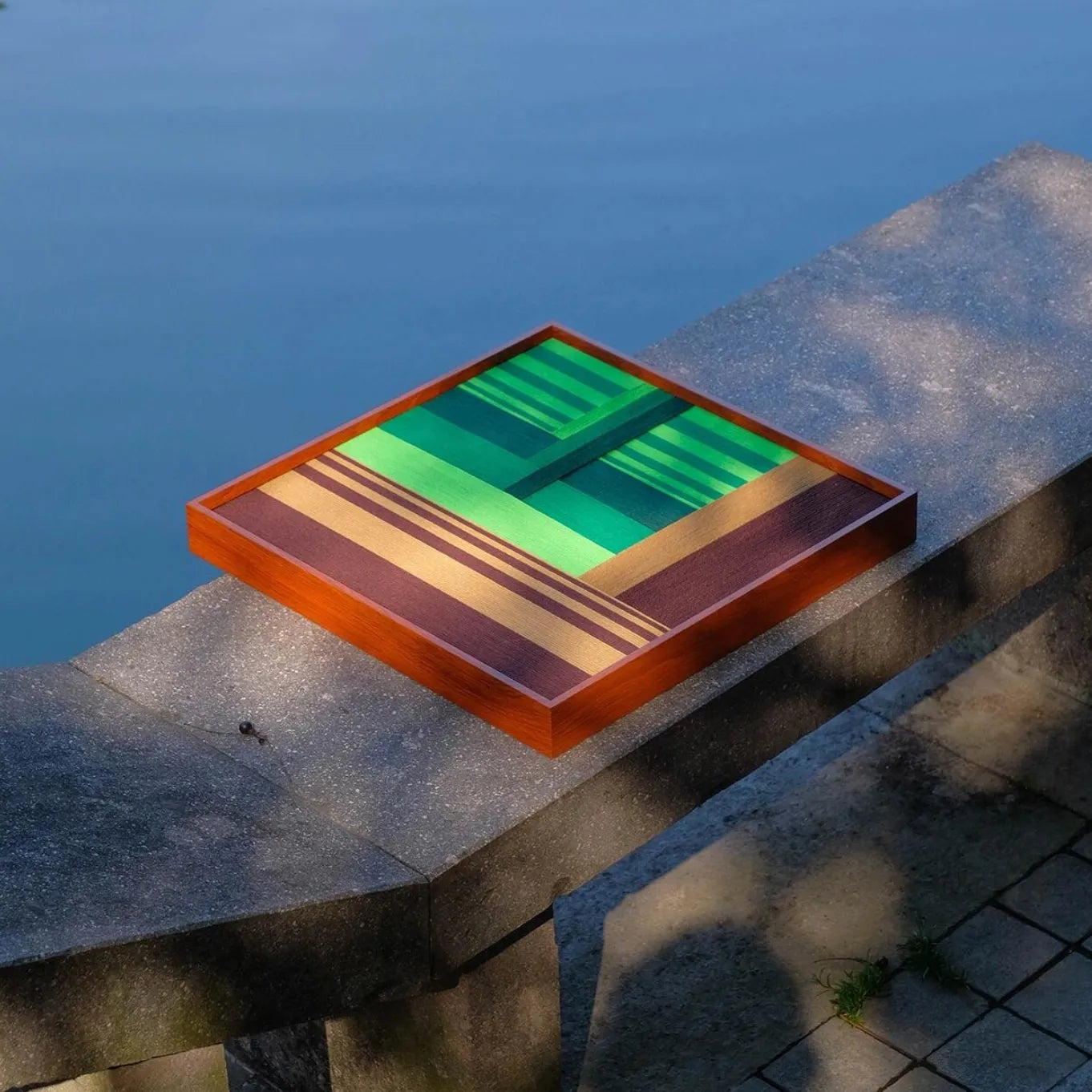 Summer Mountain Walk yarn wall art resting on a stone ledge outdoors, highlighting its deep green stripe blocks and rich wool texture.