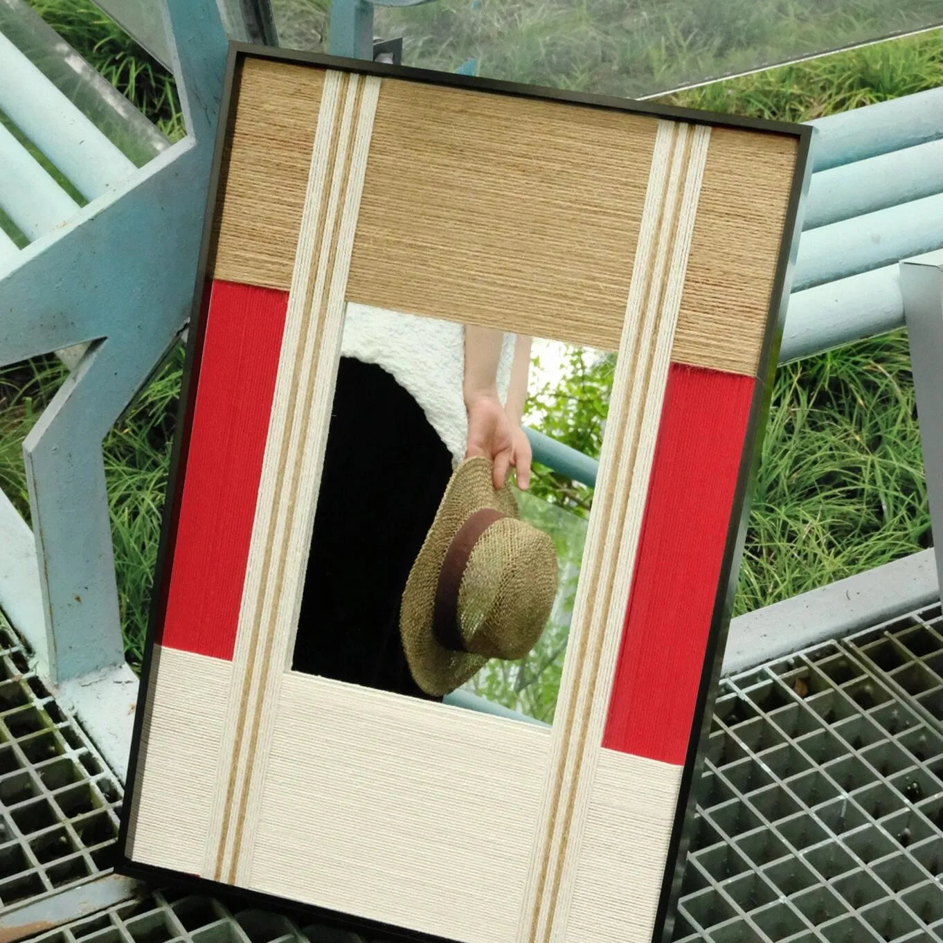 The Sun Always Rises yarn mirror art placed leaning on a metal grate, showcasing the seamless join between oatmeal and bold red wool stripe panels.