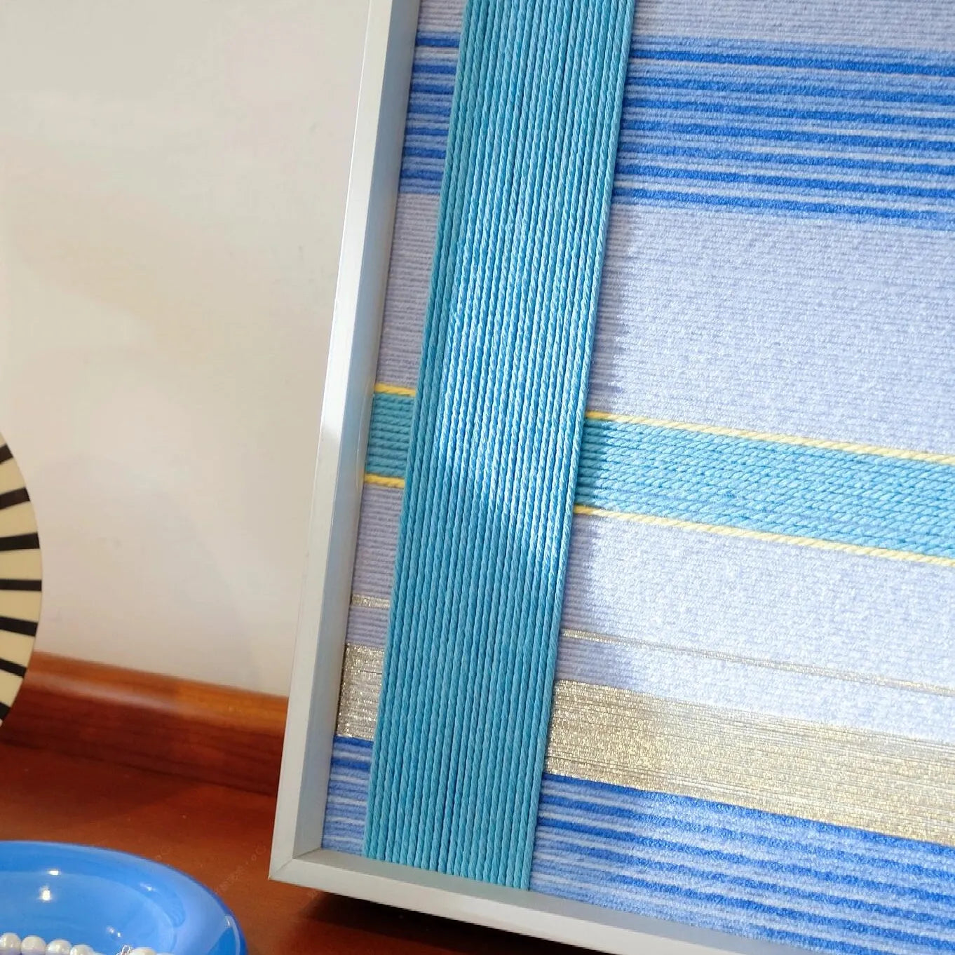 That Summer, the Blue Sea wool yarn wall art styled next to a table lamp, showcasing its crisp vertical blue stripe pattern