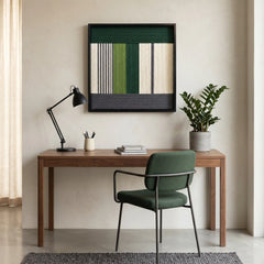 Square hand-woven textile art piece displayed above a minimalist wooden desk and green office chair in a sunlit workspace.