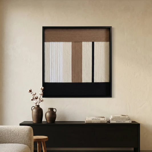 Modern hand-woven wool wall hanging displayed above a black console table in a minimalist room with warm beige plaster walls.