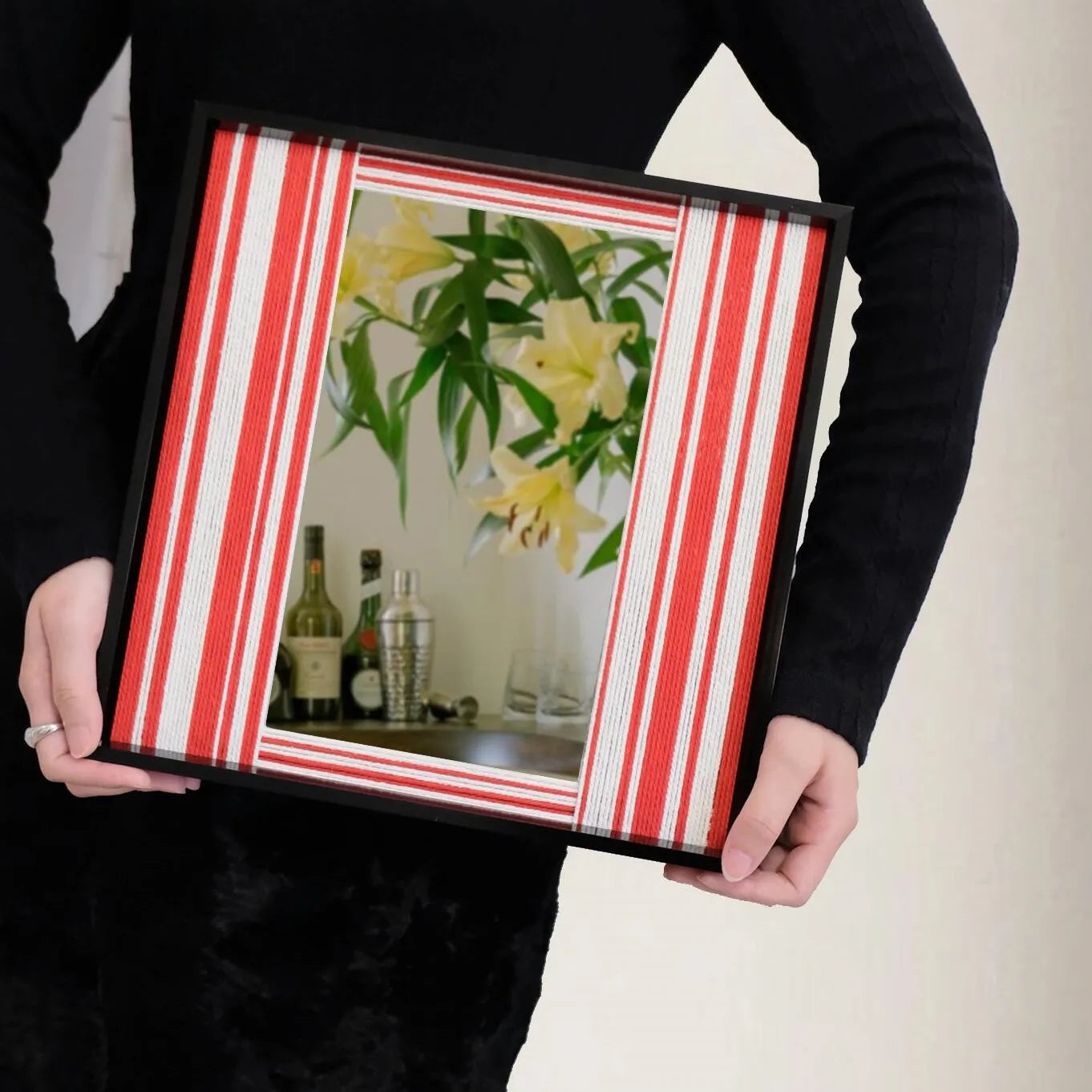 Hand-held view of Wave Tide yarn mirror art showing its compact square shape, bold red and white stripe pattern, and sturdy black frame.