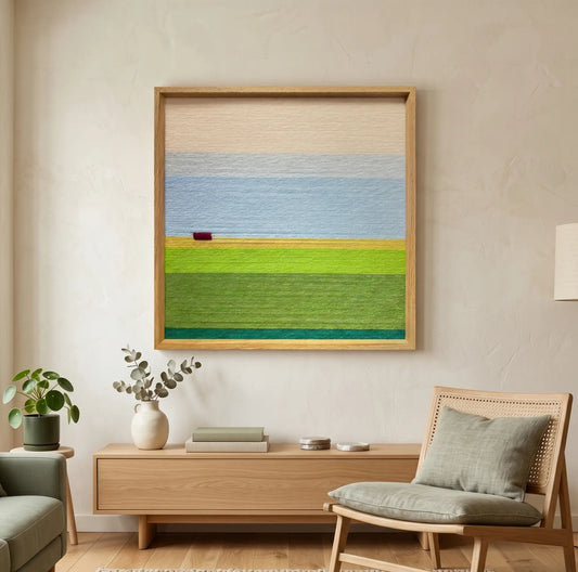 Square hand-woven wool wall art depicting a minimalist landscape with light blue sky and vibrant green fields, hanging above a modern oak sideboard and woven lounge chair.