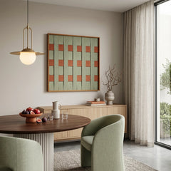 Square hand-woven textile art piece with orange and sage green accents displayed in a warm, cozy dining space with chairs.