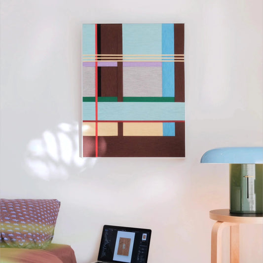 City Rhythms framed fiber wall art with a light blue, brown, red, green and lavender geometric grid hanging on a white wall beside a blue-green table lamp