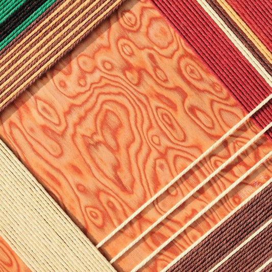 Close-up of Christmas Melody fiber wall art showing red, green, cream and brown cord lines surrounding a warm wood-grain pattern